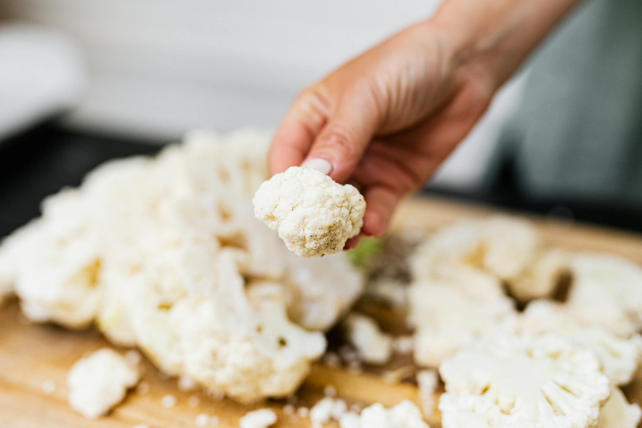 A hand holds a fresh cauliflower floret over a wooden board with scattered florets, showcasing a cooking preparation.